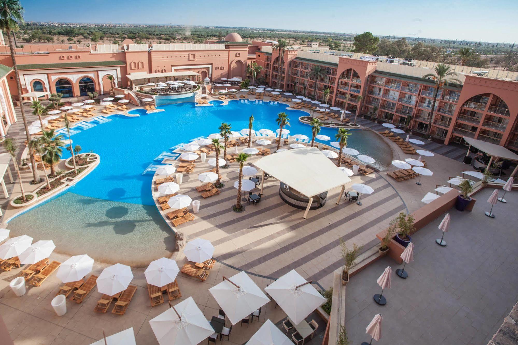 Savoy Le Grand Hotel Marrakech Марракеш Екстер'єр фото The photo shows a large, luxurious hotel resort with a spacious swimming pool at its center. The pool area features several lounge chairs and umbrellas arranged around the water, providing a relaxing atmosphere for guests. Surrounding the pool are la